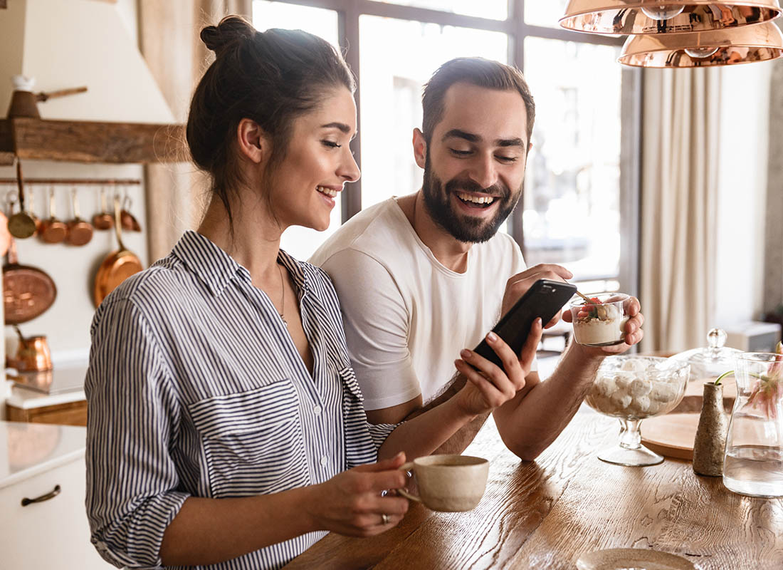 Read-Our-Reviews-Young-Couple-Smiles-as-They-Use-a-Smartphone-in-Their-Bright-Kitchen-Enjoying-Coffee